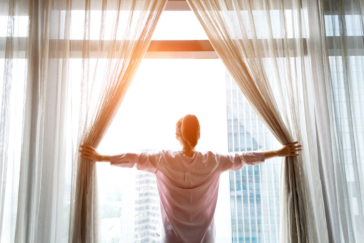 A man opening up curtains, looking out the window to bright sunlight.